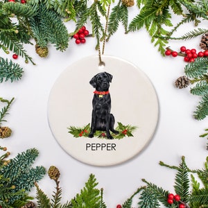 May include: A white ceramic Christmas ornament featuring a black Labrador Retriever sitting with a red collar and the name "Pepper" written below.