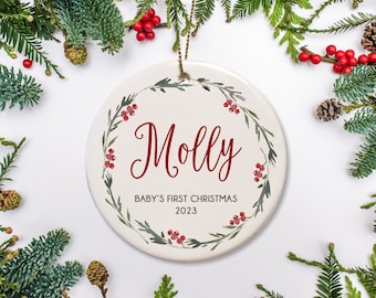Puede incluir: Adorno navideño de cerámica blanca con un diseño de corona verde y bayas rojas. El adorno tiene el nombre "Molly" y el texto "Baby's First Christmas 2023" escrito en él.