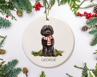 Peut inclure: Une boule de Noël ronde en céramique blanche avec un chien noir portant une écharpe rouge et blanche à rayures. Le chien est assis sur un lit de feuilles vertes et de baies rouges. La boule est inscrite du nom "Georgie" en bas.