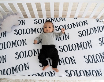 May include: A white crib sheet with the name "Solomon" printed in black all over. A baby is lying on the sheet wearing a grey long-sleeved shirt and black pants.