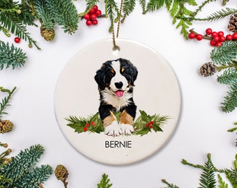 May include: A white ceramic Christmas ornament featuring a Bernese Mountain Dog puppy with black, white, and brown fur. The puppy is sitting with its tongue sticking out and surrounded by green holly leaves and red berries. The ornament is personalized with the name "Bernie".
