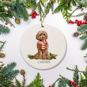 May include: A round ceramic ornament featuring a brown and white dog wearing a red and white striped scarf. The ornament is decorated with green holly and red berries. The text "CARMEN" is printed on the ornament.