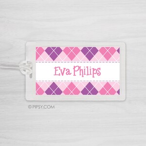 May include: A pink and purple argyle patterned luggage tag with the name "Eva Phillips" printed in pink on a white background. The tag is made of clear plastic and has a hole for a string.