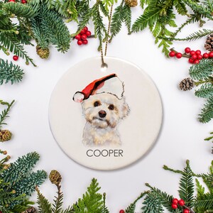 May include: White ceramic Christmas ornament with a watercolor painting of a small white dog wearing a red Santa hat. The dog's name, "COOPER", is written in black below the dog.