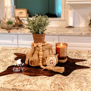 May include: A brown cowhide rug with a wooden pedestal, a potted plant, a candle, and a wooden bead garland. The pedestal has a letter "G" on it. The rug has the text "Little C Cowhides" on it.