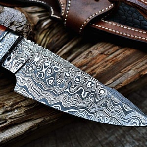May include: A close-up of a hunting knife with a patterned steel blade and a wooden handle. The knife is resting on a piece of wood.
