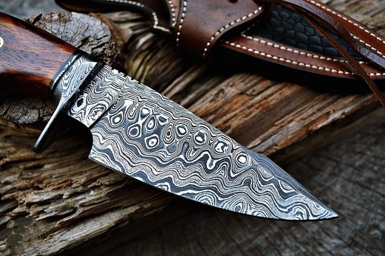 May include: A close-up of a hunting knife with a patterned steel blade and a wooden handle. The knife is resting on a piece of wood.