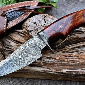 May include: A hunting knife with a patterned steel blade and a dark wood handle. The knife is resting on a piece of weathered wood.