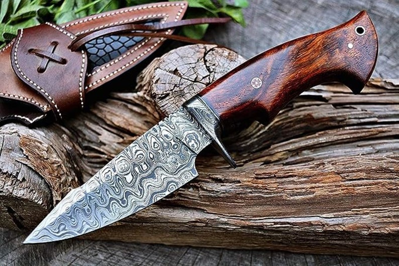 May include: A hunting knife with a patterned steel blade and a dark wood handle. The knife is resting on a piece of weathered wood.