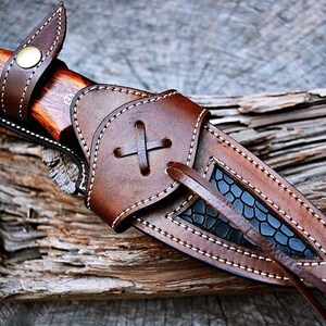May include: A brown leather knife sheath with a black patterned insert and a white stitching detail. The sheath has a strap with a cross-shaped knot.