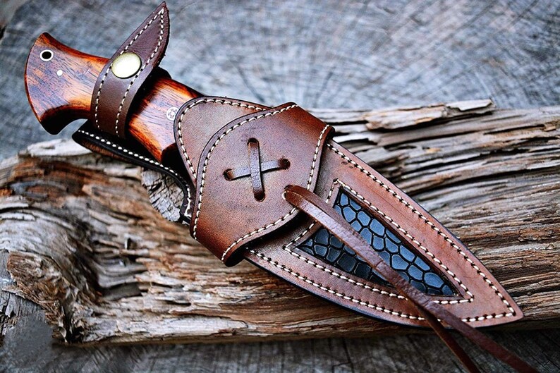 May include: A brown leather knife sheath with a black patterned insert and a white stitching detail. The sheath has a strap with a cross-shaped knot.