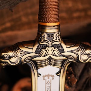 May include: Close-up of a gold-colored sword with intricate details. The hilt features a dragon-like design with a textured, brown grip.
