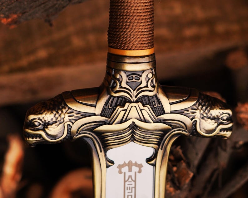 May include: Close-up of a gold-colored sword with intricate details. The hilt features a dragon-like design with a textured, brown grip.