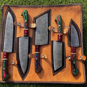 May include: A set of five kitchen knives with Damascus steel blades and multi-colored wood handles in green, red, and blue, each secured in a brown leather roll with metal snap fasteners, arranged in a row on a flat surface.