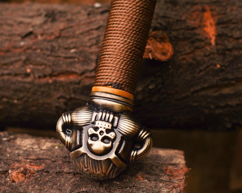 May include: Close-up of a sword pommel with a detailed skull design. The pommel is made of metal and has a brown leather wrap.