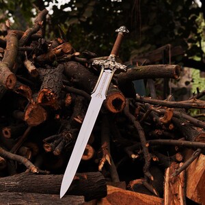 May include: A silver sword with a gold hilt and a wooden handle lies on a pile of brown branches.