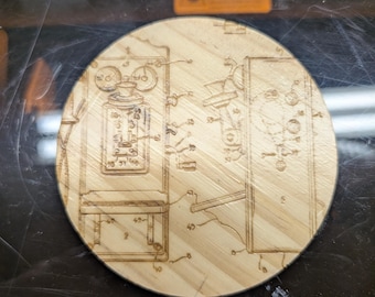 May include: A round wooden coaster with an etched diagram of a vintage telephone. The diagram includes detailed lines and numbers, showcasing the internal components of the phone. The coaster is made of light-colored wood.
