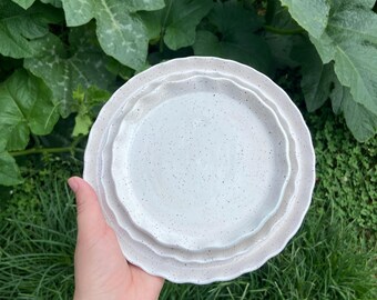 May include: A stack of three speckled white ceramic plates with a wavy rim. The plates are held in a hand, with green foliage in the background. The plates have a rustic, handmade appearance.