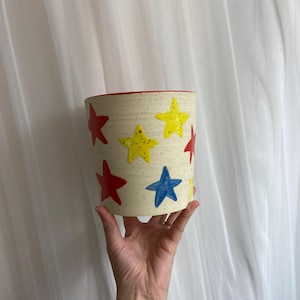 May include: A cylindrical ceramic planter with a cream-colored speckled finish. The planter is adorned with red, yellow, and blue star shapes. The interior rim is red. The planter is held by a hand.