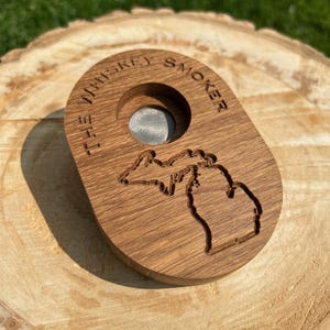 May include: A wooden whiskey smoker with the text "THE WHISKEY SMOKER" engraved around a central hole. The smoker features a detailed outline of the state of Michigan. The wood has a natural grain and is set on a wooden surface.