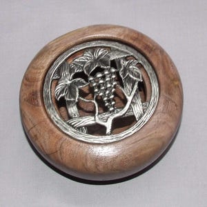 May include: A round, wooden bottle stopper with a silver-colored grape vine design. The stopper has a light brown color and a smooth finish. The grape vine design is in the center of the stopper.