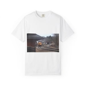 Peut inclure: T-shirt blanc avec une photographie d'un train arrivant en gare. L'image montre le train sur les rails, une gare avec un toit marron et des arbres en arrière-plan. Le train est éclairé par ses phares.