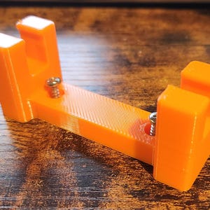 May include: Bright orange 3D-printed object with a U-shaped design. The object has two vertical rectangular supports and a horizontal base, secured with silver screws. The item is likely a bracket or holder.