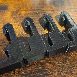 May include: Four black plastic hooks, designed to hang items on a wall or other surface. The hooks are arranged in a row, with the tips of the hooks pointing towards the right.