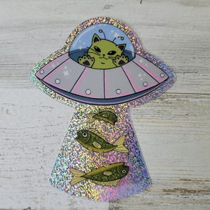 May include: A holographic sticker featuring a cartoon cat piloting a pink and gray UFO. Below the UFO are three green fish. The cat has green fur and is waving. The background is a light blue sky with stars.