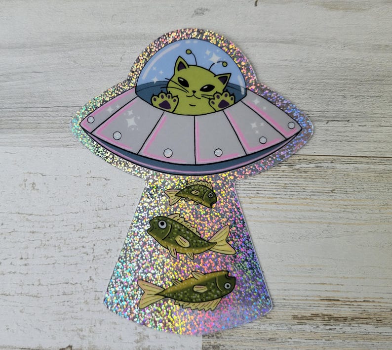 May include: A holographic sticker featuring a cartoon cat piloting a pink and gray UFO. Below the UFO are three green fish. The cat has green fur and is waving. The background is a light blue sky with stars.