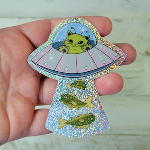 May include: A holographic sticker featuring a green cat piloting a UFO. The UFO has a light blue dome and pink accents. Below the UFO are three green fish. The sticker has a glittery, iridescent finish.
