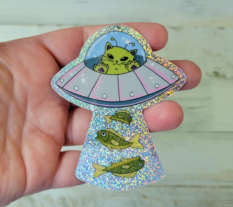 May include: A holographic sticker featuring a green cat piloting a UFO. The UFO has a light blue dome and pink accents. Below the UFO are three green fish. The sticker has a glittery, iridescent finish.
