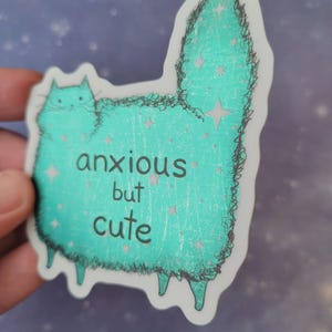 以下が含まれることがあります： 「anxious but cute」の文字が入った、ターコイズ色の猫のイラストが描かれたステッカー。猫は星のアクセントと黒いアウトラインを持つふわふわの体をしている。ステッカーは手で持たれている。