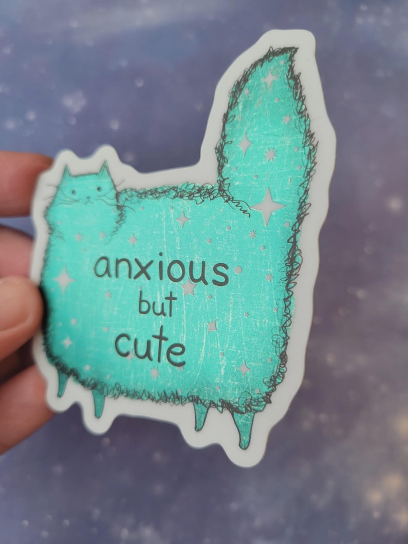 以下が含まれることがあります： 「anxious but cute」の文字が入った、ターコイズ色の猫のイラストが描かれたステッカー。猫は星のアクセントと黒いアウトラインを持つふわふわの体をしている。ステッカーは手で持たれている。