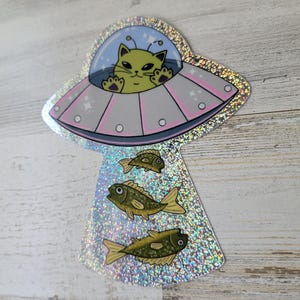 May include: A holographic sticker featuring a cartoon cat piloting a pink and silver UFO. The cat is yellow with black eyes and is surrounded by a blue bubble. Below the UFO are three green and yellow fish.