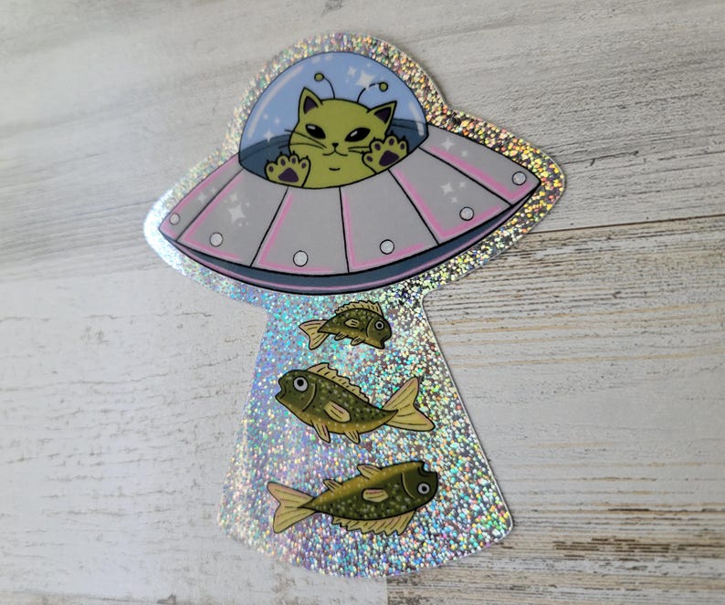 May include: A holographic sticker featuring a cartoon cat piloting a pink and silver UFO. The cat is yellow with black eyes and is surrounded by a blue bubble. Below the UFO are three green and yellow fish.