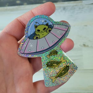 May include: A holographic sticker featuring a green cat piloting a UFO, with three yellow fish below. The UFO has a light blue dome and pink accents. The sticker has a glittery, iridescent finish.