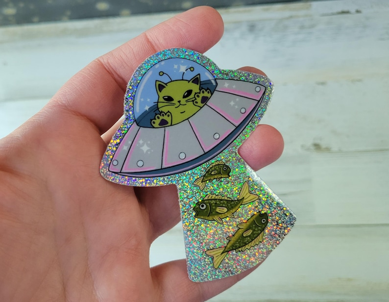 May include: A holographic sticker featuring a green cat piloting a UFO, with three yellow fish below. The UFO has a light blue dome and pink accents. The sticker has a glittery, iridescent finish.