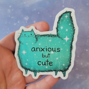 以下が含まれることがあります： 「anxious but cute」の文字が入った、ターコイズ色の猫のイラストのステッカー。猫は星のアクセントが付いたふわふわの体をしている。ステッカーは白い縁取りがあり、手で持たれている。