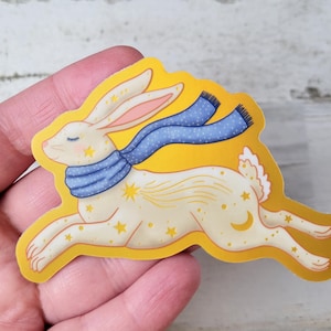 以下が含まれることがあります： 青いスカーフと金色の星を身につけたクリーム色のウサギのステッカー。ウサギは黄色の背景で走っています。ステッカーは光沢仕上げで、手で持たれています。