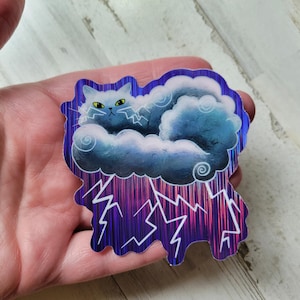 May include: A sticker featuring a cat nestled in a cloud, with lightning bolts below. The cat has yellow eyes and is colored in shades of blue and white. The sticker has a holographic, iridescent finish.
