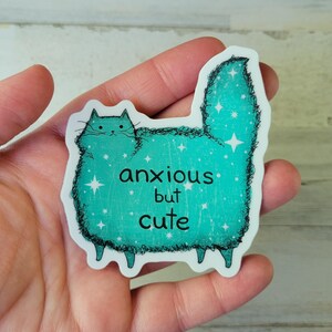 以下が含まれることがあります： 「anxious but cute」の文字が入った、猫の形をしたターコイズ色のステッカー。猫は星のアクセントが付いたふわふわの体をしている。ステッカーは手で持たれている。