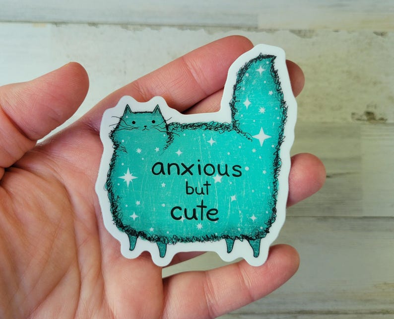 以下が含まれることがあります： 「anxious but cute」の文字が入った、猫の形をしたターコイズ色のステッカー。猫は星のアクセントが付いたふわふわの体をしている。ステッカーは手で持たれている。
