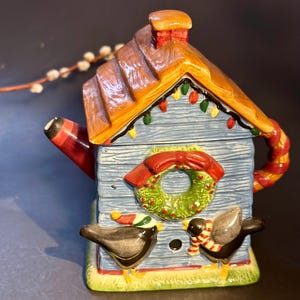 May include: A ceramic teapot shaped like a birdhouse, painted in blue, brown, red, and green. The teapot features a roof, chimney, and a wreath. Two black birds with scarves and hats are on the front. The teapot is decorated with Christmas lights.