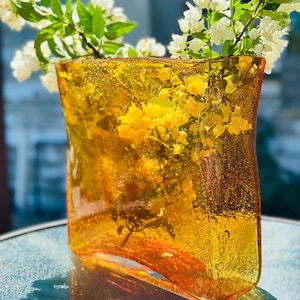May include: A square-shaped amber glass vase with a textured, bubbly surface. The vase features a curved, concave front panel and holds a bouquet of white flowers with green stems and leaves, visible through the translucent orange glass.