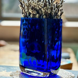 May include: A vibrant blue glass vase filled with pussy willow branches. The vase has a rectangular shape with a thick base and is sitting on a decorative doily. The pussy willow branches have soft, fuzzy buds.