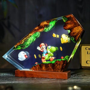 May include: A decorative lamp featuring a scene from the Super Mario video game. The lamp has a wooden base and a clear, angled resin body. Inside, Mario rides Yoshi, with coins, stars, and a ghost. The top edge has a wood and green moss design.