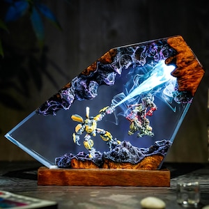 May include: A unique resin art piece featuring two robot figures in a battle scene. The artwork is encased in a clear, angled resin block with a wooden base. The figures are yellow and green, with a blue energy beam.