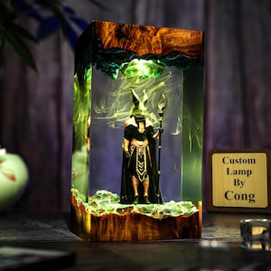 以下が含まれることがあります： 透明な樹脂の中に魔法使いのフィギュアが入った長方形のランプ。 ランプは木製のベースと上部があり、緑と黄色のアクセントがあります。 フィギュアは黒と金で、杖を持っています。 「Custom Lamp By Cong」というテキストが木製のプラークにあります。