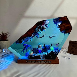 May include: A resin lamp shaped like a diamond with a blue and green underwater scene inside. The scene includes a sunken ship, an octopus, a turtle, and coral. The lamp is lit from the bottom, casting a blue glow. The lamp is on a wooden base with the text "CustomLampByCong" engraved on it.
